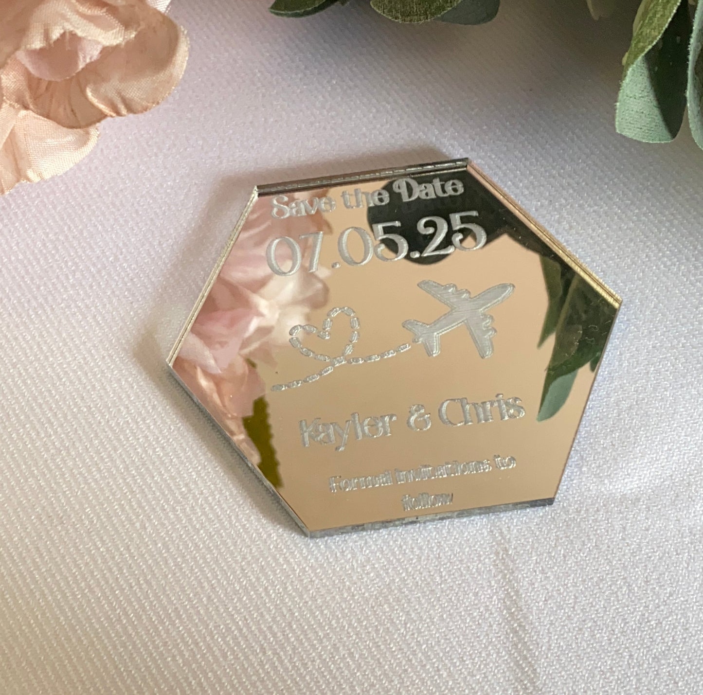 A hexagonal Destination Wedding Save The Date Magnet in mirror silver.The save the date has the wedding date and an aeroplane with a heart shaped trail behind engraved in to the acrylic.