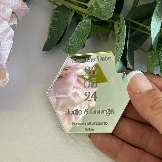 Hexagonal wedding save the date card held by a hand with greenery and flowers in the background.
The save the date is made from mirror silver acrylic with the details engraved.