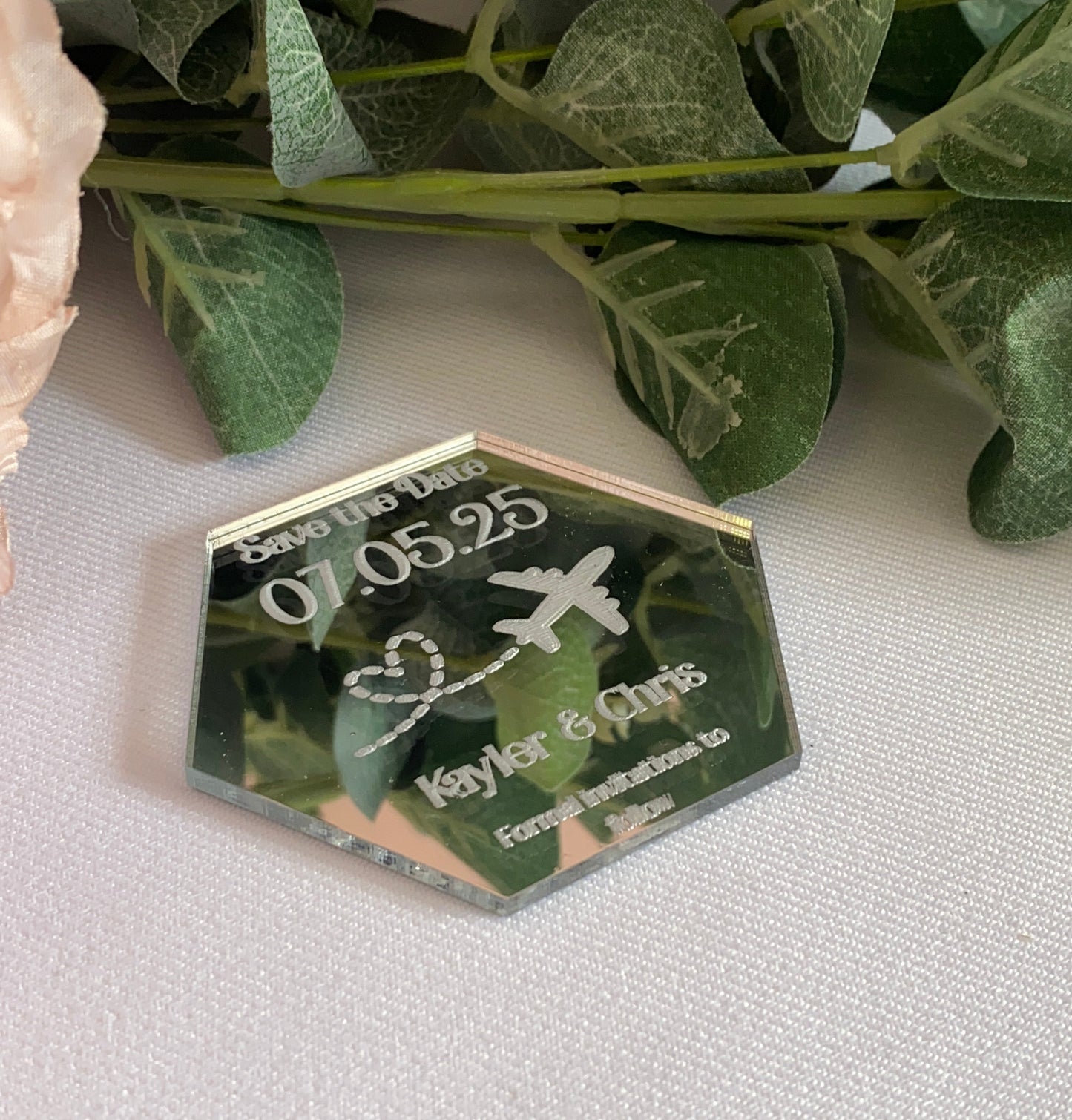 A hexagonal Destination Wedding Save The Date Magnet in mirror silver.The save the date has the wedding date and an aeroplane with a heart shaped trail behind engraved in to the acrylic.
