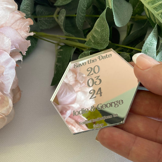 Hexagonal wedding save the date card held by a hand with greenery and flowers in the background.
The save the date is made from mirror silver acrylic with the details engraved.